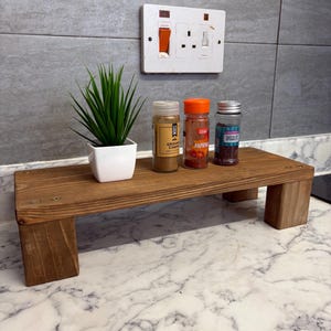 May include: A wooden spice rack with three spice jars and a small potted plant. The rack is rectangular with four legs. The jars are labelled "Ground Cumin", "Paprika", and "Chocolate Sprinkles". The rack is approximately 30 cm long.