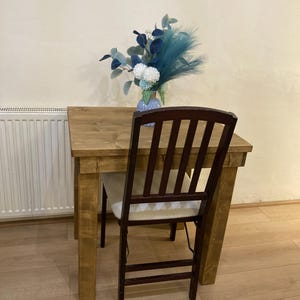K&ouml;nnte beinhalten: Ein kleiner Holztisch mit hellbrauner Oberfl&auml;che und einem einzelnen braunen Stuhl. Eine Vase mit blauen und wei&szlig;en Blumen steht auf dem Tisch.