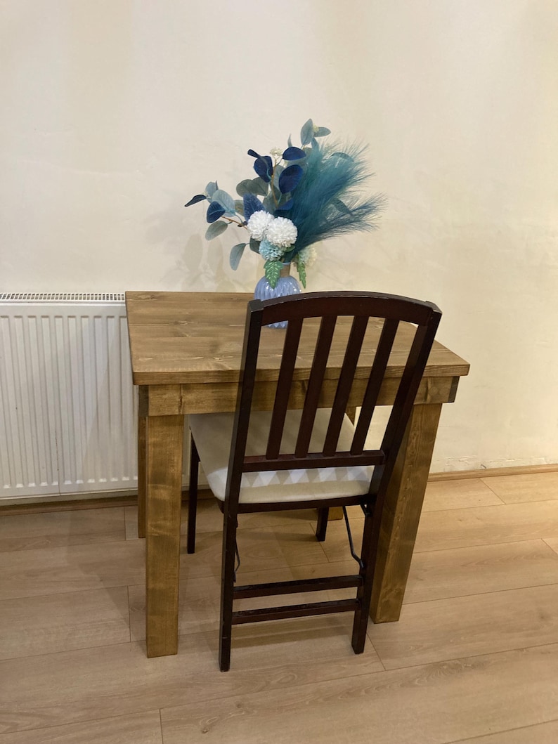 K&ouml;nnte beinhalten: Ein kleiner Holztisch mit hellbrauner Oberfl&auml;che und einem einzelnen braunen Stuhl. Eine Vase mit blauen und wei&szlig;en Blumen steht auf dem Tisch.