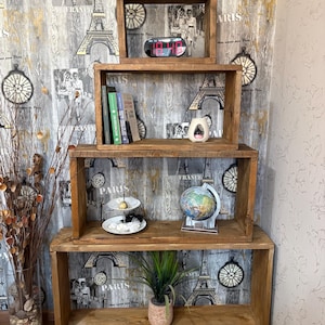 May include: A four-tier wooden shelving unit constructed from rectangular boxes of varying sizes stacked vertically. The shelves feature a medium-brown wood grain finish and contain various decorative items including books, a globe, and a clock.