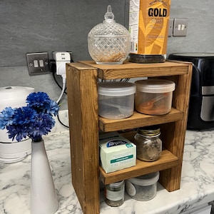 May include: A rustic, three-tiered wooden shelf unit, stained a warm brown, displaying kitchen essentials. It holds a glass jar, a coffee container, and several clear plastic containers. A white vase with blue flowers is on the counter.