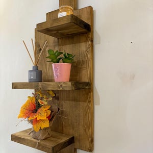 May include: A rustic, wooden wall shelf with three tiers. The shelves hold decorative items, including a small potted plant, a jar, and a bundle of fall-colored leaves. The wood has a warm, natural finish.