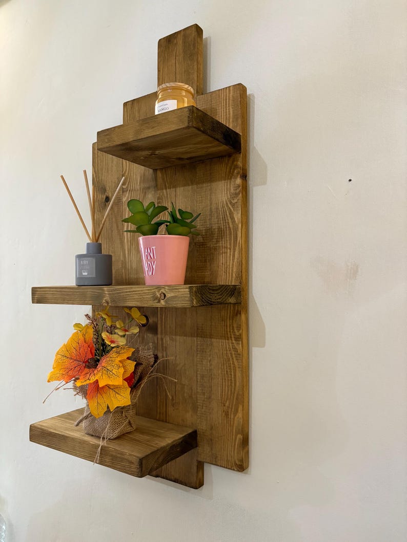 May include: A rustic, wooden wall shelf with three tiers. The shelves hold decorative items, including a small potted plant, a jar, and a bundle of fall-colored leaves. The wood has a warm, natural finish.