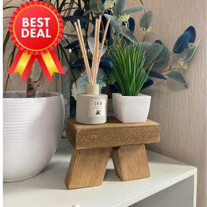 May include: A small wooden stool with a light brown finish. The stool has two legs on each side and is decorated with a diffuser bottle and a potted plant. The diffuser bottle is grey and has the word "CALM" printed on it. The potted plant is in a white pot and has green leaves.