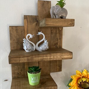 May include: A rustic, wooden wall shelf unit with three shelves. The shelves hold decorative items, including two swan figurines, a small elephant planter, and a green pot with the text "WILL SURVIVE".