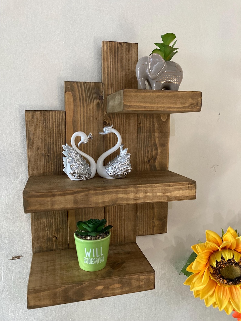 May include: A rustic, wooden wall shelf unit with three shelves. The shelves hold decorative items, including two swan figurines, a small elephant planter, and a green pot with the text "WILL SURVIVE".