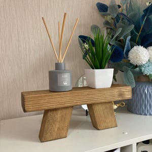 Könnte beinhalten: Ein Holzregal mit zwei Beinen, auf dem sich eine graue Diffuserflasche mit Schilfrohren und eine kleine Topfpflanze befinden. Das Regal steht auf einem weißen Regal mit einer weißen Tasche mit der Aufschrift "LUNGO" und einer Flasche Lotion mit der Aufschrift "NEW Etin Skin Solution Lotion" im Hintergrund.