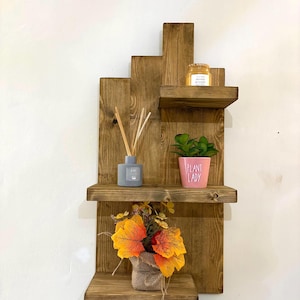 May include: A rustic, wooden wall shelf with three tiers. The shelves hold a small potted plant, a jar of honey, a gray diffuser, and a burlap-wrapped floral arrangement with orange and yellow leaves. The shelf is made of stained wood.