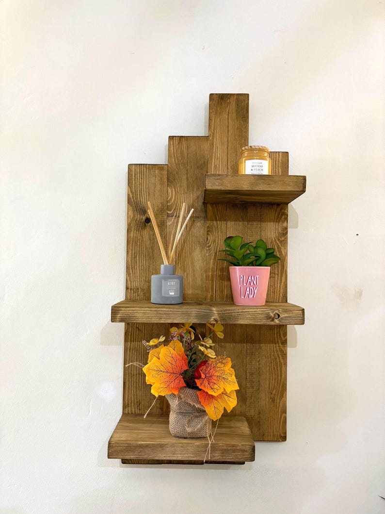 May include: A rustic, wooden wall shelf with three tiers. The shelves hold a small potted plant, a jar of honey, a gray diffuser, and a burlap-wrapped floral arrangement with orange and yellow leaves. The shelf is made of stained wood.