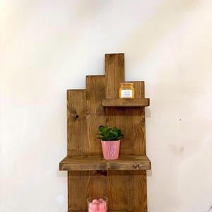 May include: A rustic, wooden wall shelf with three tiers. The shelves hold a small potted plant, a jar, and a candle. The pink pot has the words "PLANT LADY" printed on it. The shelf is made of stained wood.
