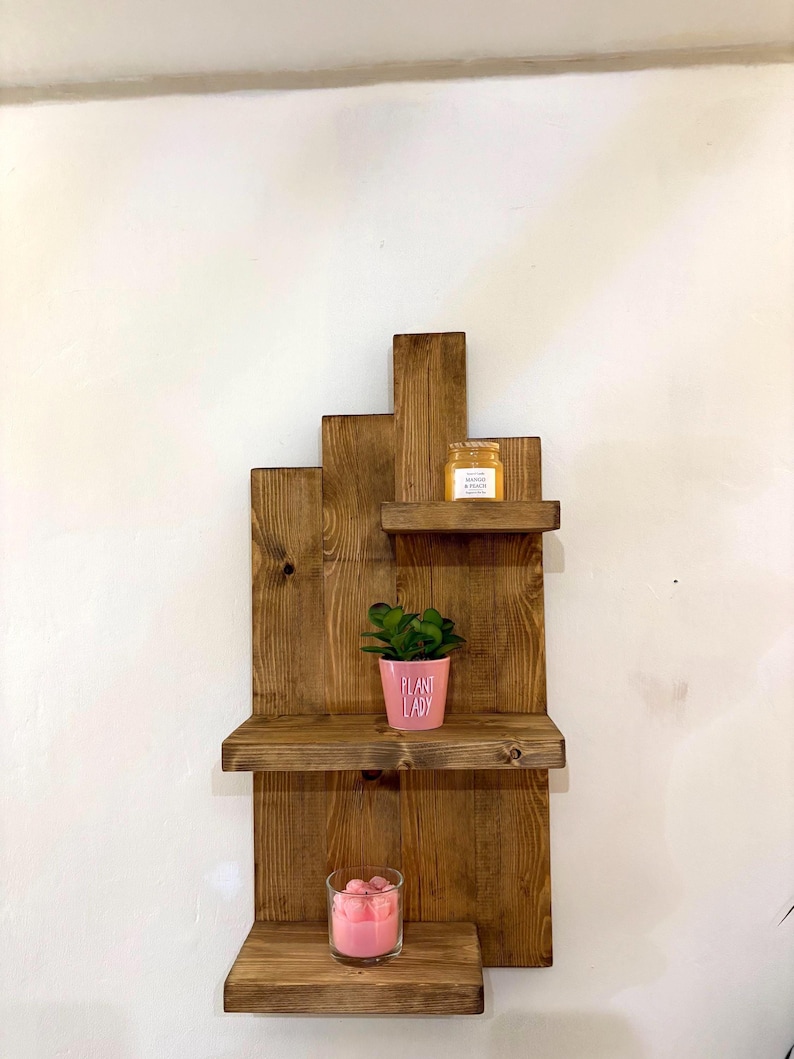 May include: A rustic, wooden wall shelf with three tiers. The shelves hold a small potted plant, a jar, and a candle. The pink pot has the words "PLANT LADY" printed on it. The shelf is made of stained wood.
