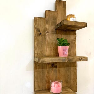 May include: A rustic, wooden wall shelf with three tiers. The shelves are stained a warm brown and hold a small potted plant, a candle in a glass, and a jar. The pink pot has the words "PLANT LADY" printed on it.