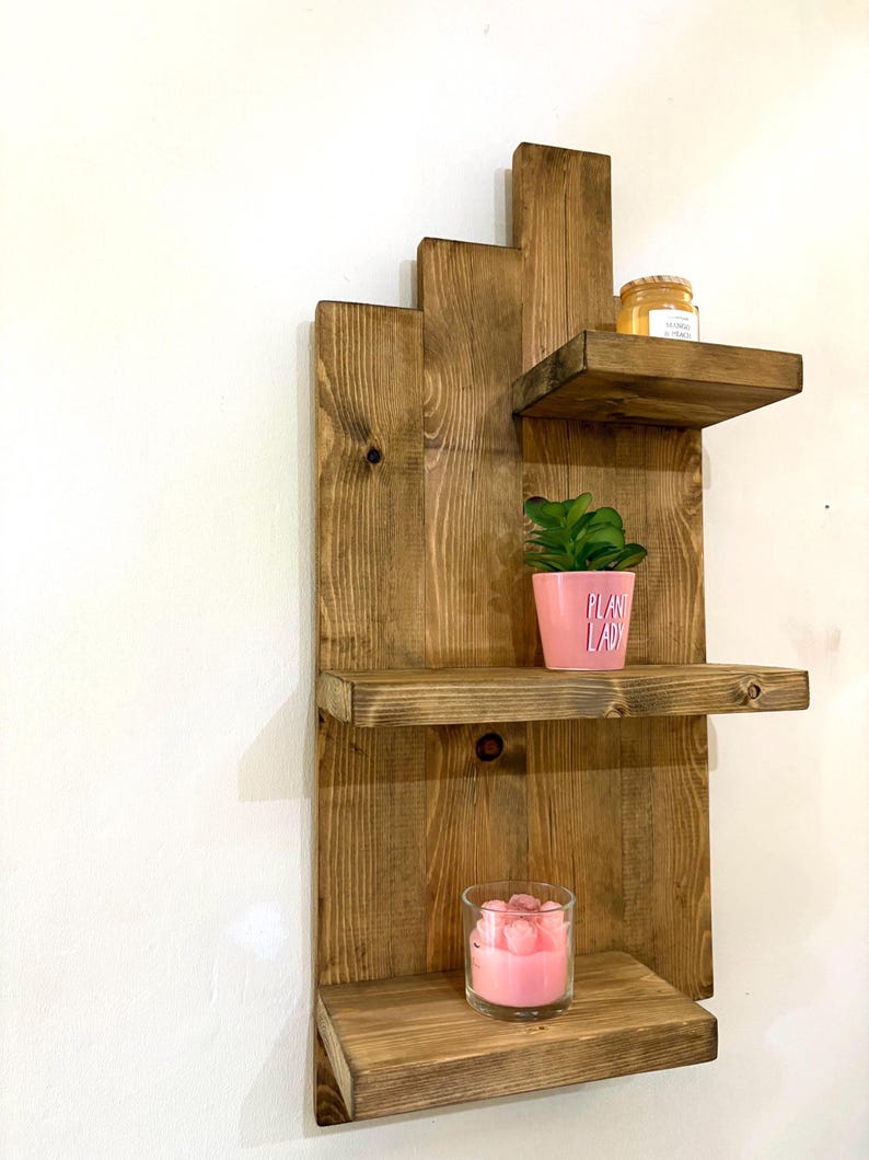 May include: A rustic, wooden wall shelf with three tiers. The shelves are stained a warm brown and hold a small potted plant, a candle in a glass, and a jar. The pink pot has the words "PLANT LADY" printed on it.