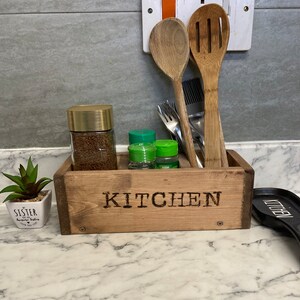 May include: A rustic wooden kitchen organiser with the word "KITCHEN" branded on the front. It holds a jar of coffee, spice jars, wooden spoons, and other utensils. A small potted plant is to the left.