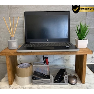 May include: A stained wooden laptop stand supporting a black laptop. A small white square pot with a green plant is to the right. Below, a desk organiser, tape, and a computer mouse are visible.