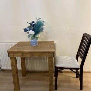 May include: A small wooden table with a light brown finish and a white padded chair. A blue and white vase with blue flowers sits on the table.