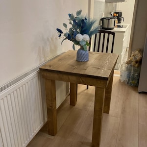 K&ouml;nnte beinhalten: Ein kleiner Holztisch mit hellbrauner Oberfl&auml;che. Der Tisch hat vier Beine und eine quadratische Tischplatte. Eine blaue und wei&szlig;e Blumenarrangements steht auf dem Tisch in einer blauen Vase.