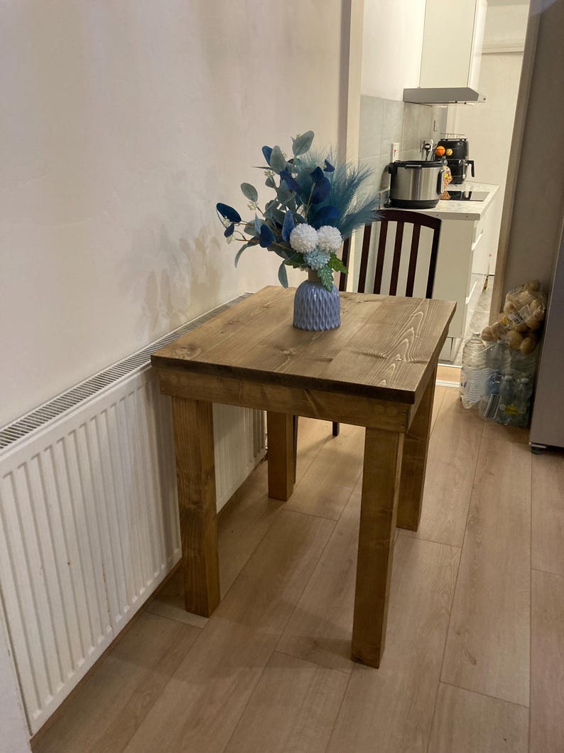 K&ouml;nnte beinhalten: Ein kleiner Holztisch mit hellbrauner Oberfl&auml;che. Der Tisch hat vier Beine und eine quadratische Tischplatte. Eine blaue und wei&szlig;e Blumenarrangements steht auf dem Tisch in einer blauen Vase.