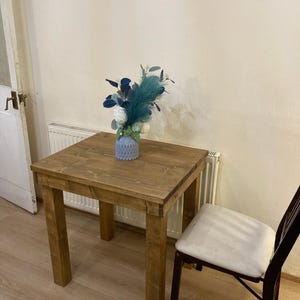 K&ouml;nnte beinhalten: Ein kleiner Holztisch mit hellbrauner Oberfl&auml;che. Der Tisch hat eine quadratische Tischplatte und vier Beine. Eine blaue Vase mit einem Blumenarrangement steht auf dem Tisch. Ein wei&szlig;er Stuhl steht neben dem Tisch.