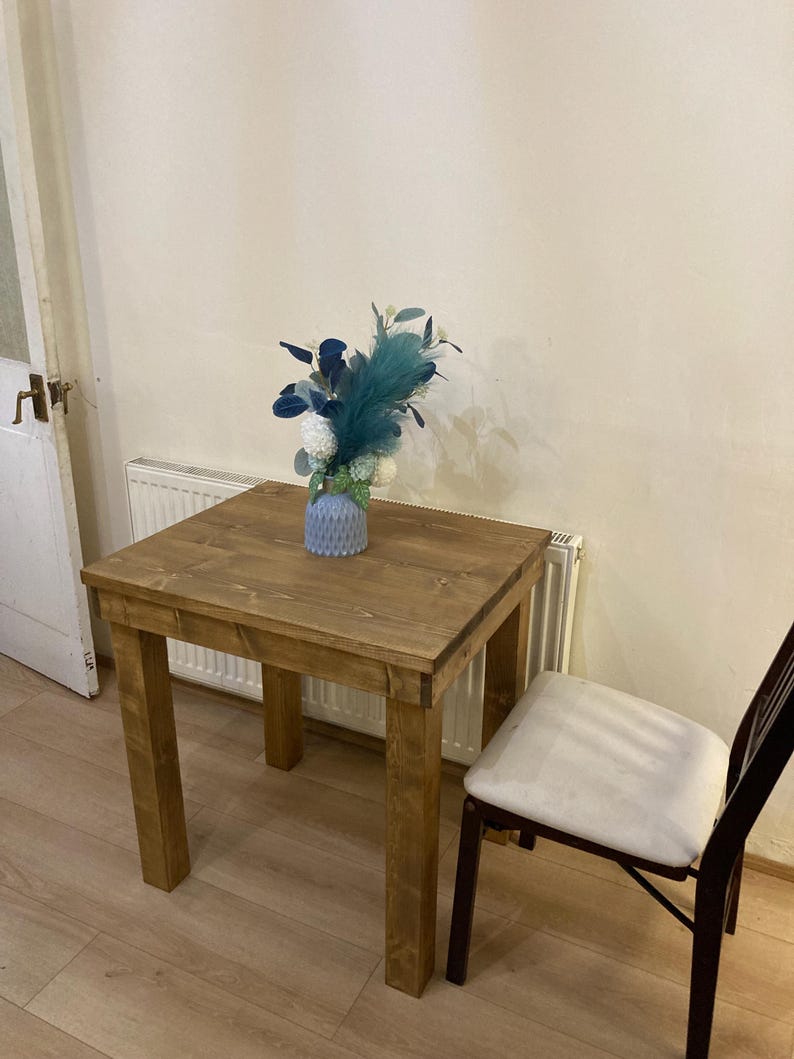 K&ouml;nnte beinhalten: Ein kleiner Holztisch mit hellbrauner Oberfl&auml;che. Der Tisch hat eine quadratische Tischplatte und vier Beine. Eine blaue Vase mit einem Blumenarrangement steht auf dem Tisch. Ein wei&szlig;er Stuhl steht neben dem Tisch.