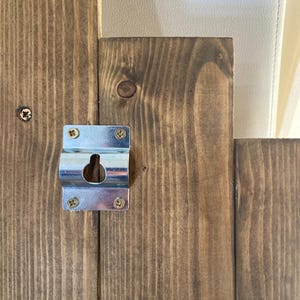 May include: Close-up of a wooden panel with a metal bracket attached. The wood has a dark brown stain and visible grain. The bracket is silver and secured with four screws, featuring a keyhole for hanging.