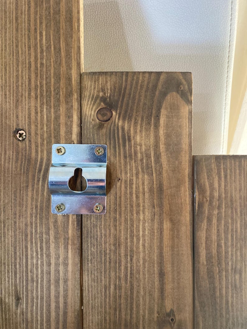 May include: Close-up of a wooden panel with a metal bracket attached. The wood has a dark brown stain and visible grain. The bracket is silver and secured with four screws, featuring a keyhole for hanging.