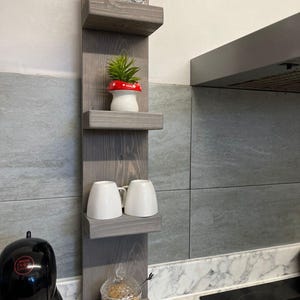 Könnte beinhalten: Ein graues Holzregal an der Wand mit vier Ablagen. Die oberste Ablage hat ein Paar weiße Keramikschwäne, die zweite Ablage hat eine kleine Topfpflanze mit einem roten Pilz, die dritte Ablage hat zwei weiße Keramikbecher und die unterste Ablage hat ein Glasgefäß mit Deckel und einen kleinen Holzlöffel.