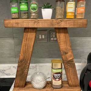 Könnte beinhalten: Ein zweistufiges Holzregal, die obere Stufe enthält sechs Gewürzgläser mit Etiketten wie "Oregano", "Petersilie", "Thymian", "Kreuzkümmel" und eine kleine Topfpflanze. Die untere Stufe enthält ein Glasgefäß und ein Glas Nescafé Gold Blend Instantkaffee.