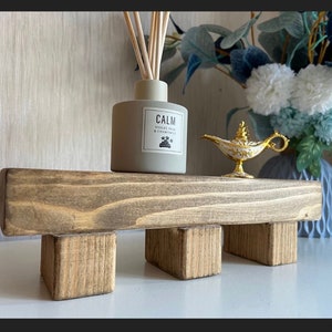 Könnte beinhalten: Ein rustikaler Holzständer mit Used-Look, auf dem ein grauer Aromatherapie-Diffusor mit der Aufschrift "CALM" und eine goldene Dschinn-Lampe stehen. Eine Vase mit blau-weißen Blumen ist ebenfalls zu sehen.