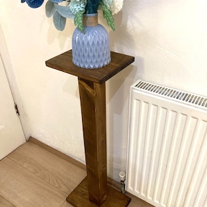 May include: A tall, dark brown wooden plant stand featuring a square top and a square base connected by a central vertical pillar. A light blue, textured, bulbous vase containing a bouquet of white and blue artificial flowers sits on top.