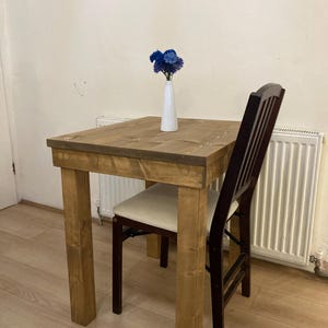 K&ouml;nnte beinhalten: Ein kleiner Holztisch mit quadratischer Tischplatte und einem Stuhl. Der Tisch ist aus Naturholz gefertigt und hat eine rustikale Oberfl&auml;che. Eine Vase mit blauen Blumen steht auf dem Tisch.