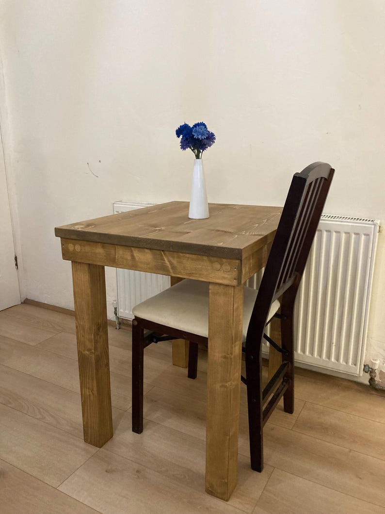K&ouml;nnte beinhalten: Ein kleiner Holztisch mit quadratischer Tischplatte und einem Stuhl. Der Tisch ist aus Naturholz gefertigt und hat eine rustikale Oberfl&auml;che. Eine Vase mit blauen Blumen steht auf dem Tisch.