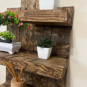 May include: A rustic, multi-tiered wooden shelf unit with a dark brown finish. The shelves display small potted plants, including a succulent, a green plant, and a plant with pink flowers. A glass diffuser bottle with reeds is also visible.