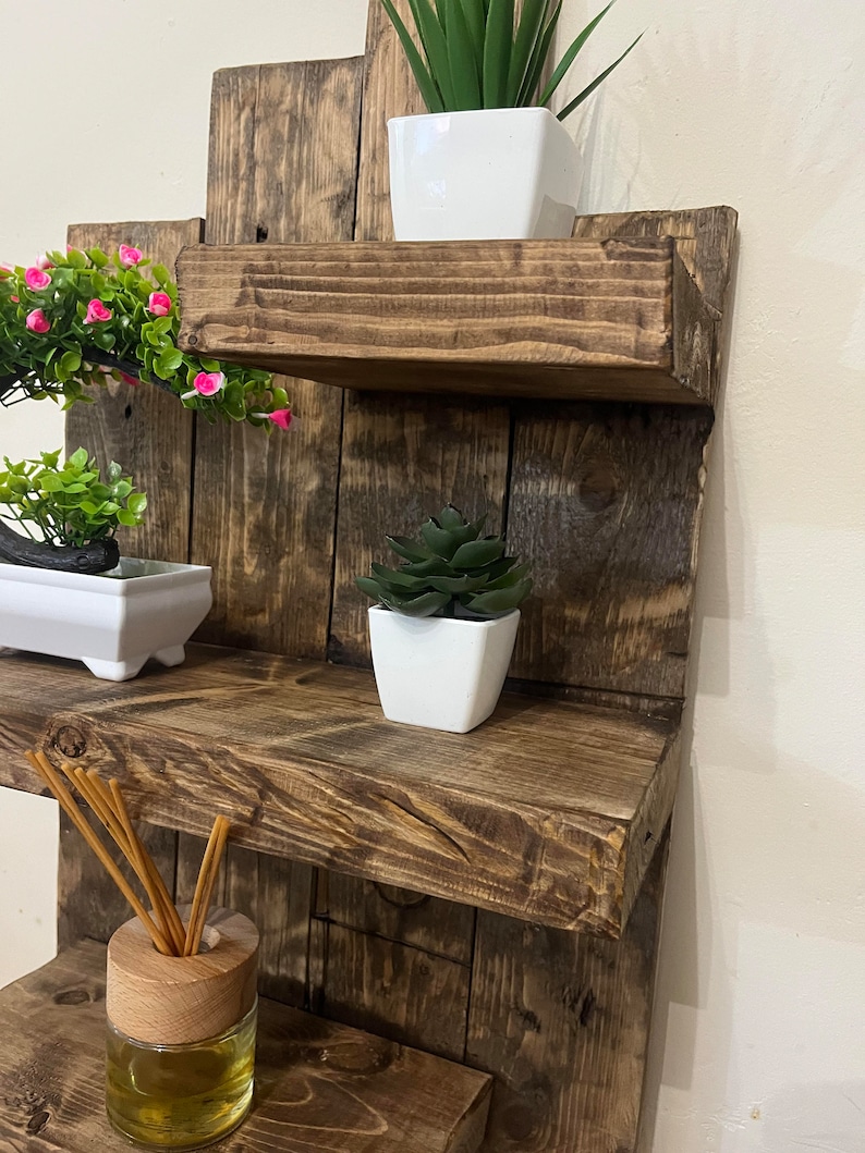 May include: A rustic, multi-tiered wooden shelf unit with a dark brown finish. The shelves display small potted plants, including a succulent, a green plant, and a plant with pink flowers. A glass diffuser bottle with reeds is also visible.