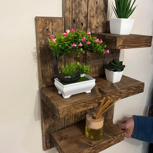 May include: A rustic, wooden wall shelf with three tiers, stained a dark brown. The shelves hold small potted plants, including a bonsai tree with pink flowers, and a reed diffuser. The shelf is approximately 24 inches wide.