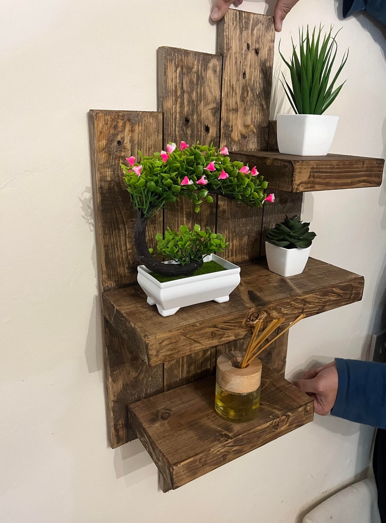 May include: A rustic, wooden wall shelf with three tiers, stained a dark brown. The shelves hold small potted plants, including a bonsai tree with pink flowers, and a reed diffuser. The shelf is approximately 24 inches wide.