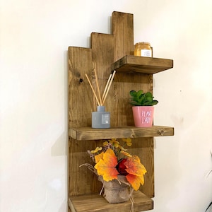 May include: A rustic, wooden wall shelf with three tiers. The shelf is stained a warm brown and features a stepped design. It holds a small potted plant, a candle, a diffuser, and a burlap-wrapped floral arrangement with orange and yellow leaves.