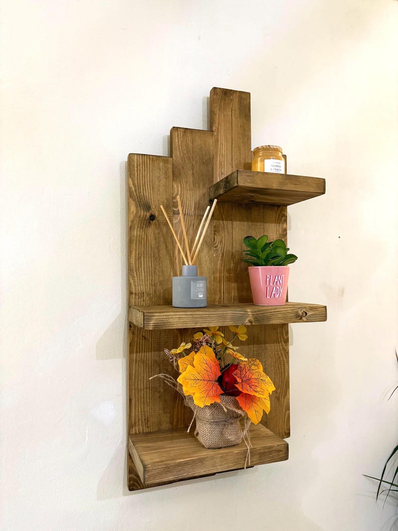May include: A rustic, wooden wall shelf with three tiers. The shelf is stained a warm brown and features a stepped design. It holds a small potted plant, a candle, a diffuser, and a burlap-wrapped floral arrangement with orange and yellow leaves.