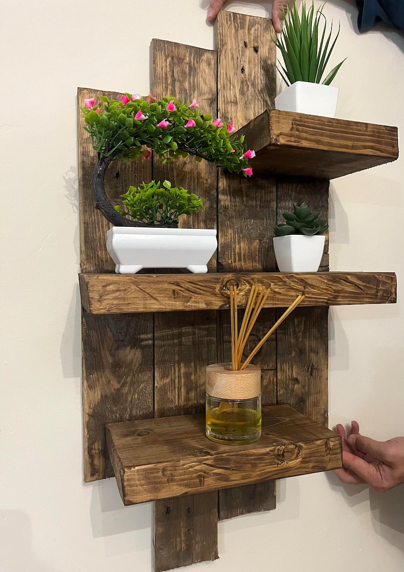May include: A rustic, wooden wall shelf with three shelves. The shelves hold small potted plants and a glass jar with reeds. The wood has a dark, weathered finish, and the plants add a touch of green and pink.