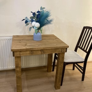 K&ouml;nnte beinhalten: Ein rustikaler Holztisch mit hellbrauner Oberfl&auml;che. Der Tisch hat eine quadratische Tischplatte und vier Beine. Eine Vase mit blauen und wei&szlig;en Blumen steht auf dem Tisch. Ein Holzstuhl mit einem wei&szlig;en Kissen steht neben dem Tisch.