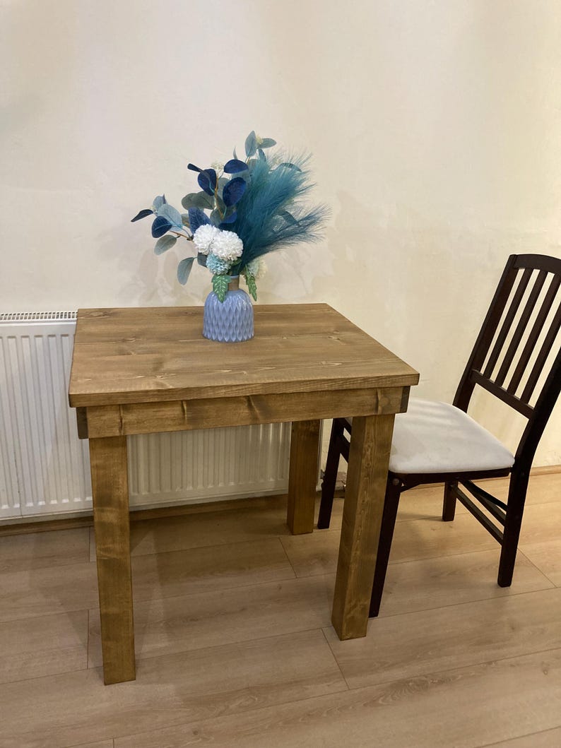 K&ouml;nnte beinhalten: Ein rustikaler Holztisch mit hellbrauner Oberfl&auml;che. Der Tisch hat eine quadratische Tischplatte und vier Beine. Eine Vase mit blauen und wei&szlig;en Blumen steht auf dem Tisch. Ein Holzstuhl mit einem wei&szlig;en Kissen steht neben dem Tisch.
