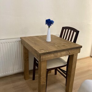 K&ouml;nnte beinhalten: Ein kleiner Holztisch mit hellbrauner Oberfl&auml;che und einer wei&szlig;en Vase mit blauen Blumen darauf. Ein brauner Stuhl mit wei&szlig;em Kissen ist an den Tisch gezogen.
