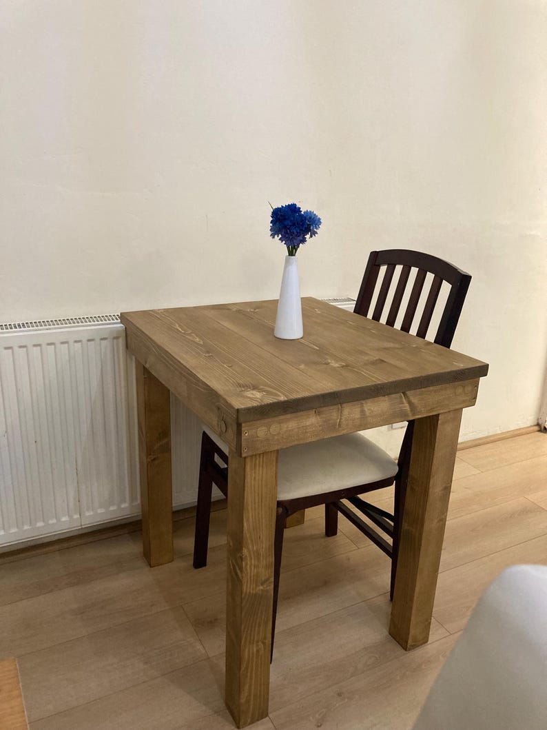 K&ouml;nnte beinhalten: Ein kleiner Holztisch mit hellbrauner Oberfl&auml;che und einer wei&szlig;en Vase mit blauen Blumen darauf. Ein brauner Stuhl mit wei&szlig;em Kissen ist an den Tisch gezogen.