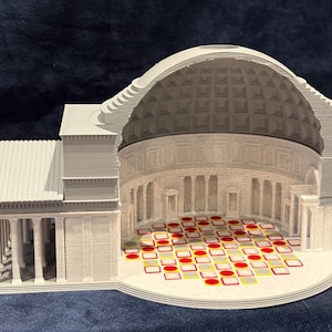 May include: A 3D-printed model of the Pantheon, a Roman temple, showcasing a cross-section view. The model is primarily light gray with a detailed dome and columns. The floor features a pattern of red, yellow, and gray squares.