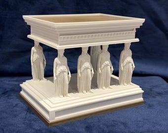 Puede incluir: Maqueta arquitectónica blanca de un templo clásico, con seis figuras femeninas como columnas que sostienen un techo rectangular. La base y el techo tienen un borde dorado. El modelo está sobre un fondo azul oscuro.