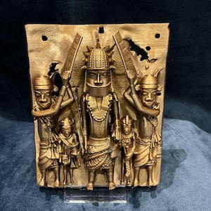May include: A rectangular bronze-coloured wall plaque featuring a central figure wearing a tall, textured headdress and neck rings, flanked by two larger figures holding rectangular objects and two smaller figures, all rendered in a relief style.