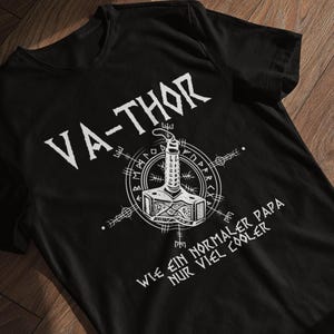 Könnte beinhalten: Schwarzes T-Shirt mit weißer Grafik von Thors Hammer und dem Text "VA-THOR" darüber. Unter dem Hammer steht der Text "WIE EIN NORMALER PAPA NUR VIEL COOLER". Das Shirt ist aus weichem Material.