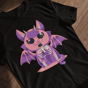 Könnte beinhalten: Schwarzes T-Shirt mit einem Cartoon-Fledermaus-Design. Die Fledermaus ist in Rosa und Lila gehalten, mit einem Skelett-Detail. Das T-Shirt hat einen Rundhalsausschnitt und kurze Ärmel. Das Design ist auf der Vorderseite zentriert.