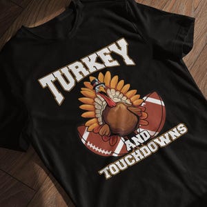 Könnte beinhalten: Schwarzes T-Shirt mit einem Thanksgiving-Motiv. Das Design zeigt einen Cartoon-Truthahn mit Zylinder, positioniert zwischen zwei American-Football-Bällen. Der Text "TURKEY AND TOUCHDOWNS" ist in Weiß dargestellt.