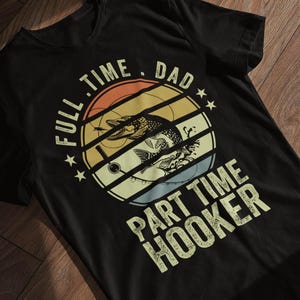 Könnte beinhalten: Schwarzes T-Shirt mit einem Retro-Design eines aus dem Wasser springenden Fisches, umgeben von dem Text "FULL TIME DAD PART TIME HOOKER". Das Design beinhaltet Sterne und eine Vintage-Farbpalette.